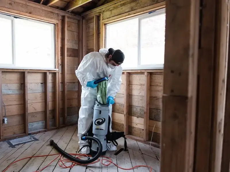 Technician in protective coveralls vacuuming a room opened to the studs