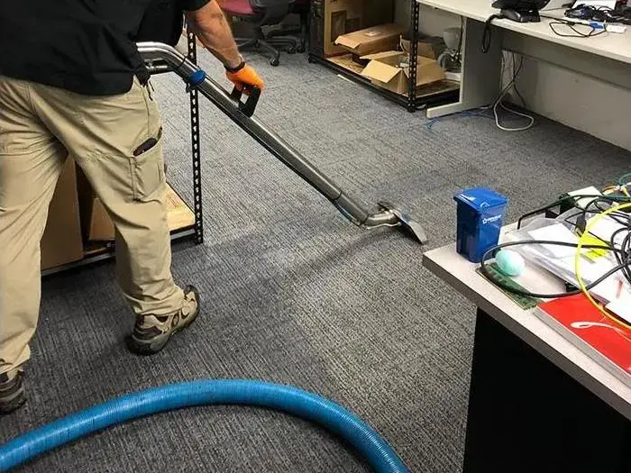 Technician running an extraction wand over wet carpet tiles in an office